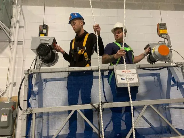 Two trainees in hard hats and harnesses on the swing stage rig